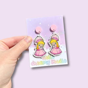 Op de afbeelding: Roze Princess Peach oorbellen met een bijpassende roze top en een witte paraplu. De oorbellen zijn bevestigd aan een lichtpaarse kaart met de tekst "hello dreamy studio".