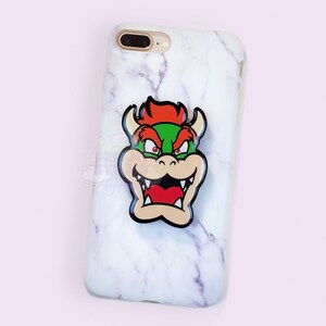 May include: A white marble iPhone case with a red, green, and black Bowser character phone grip.