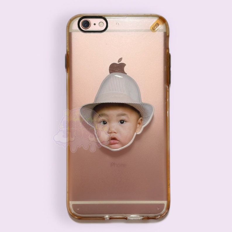 Op de afbeelding: Een ros&eacute;gouden iPhone 6 met een transparante hoes. Op de telefoon zit een sticker van een baby met een witte hoed. Het Apple-logo en de camera zijn zichtbaar. Het woord "iPhone" staat onderaan gedrukt.