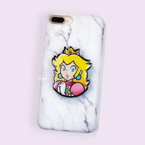 Puede incluir: Una funda de teléfono de mármol blanco con un soporte pop que presenta una imagen de dibujos animados de la princesa Peach de la serie de videojuegos Super Mario Bros. El soporte pop es negro con una imagen rosa y amarilla de la princesa Peach.