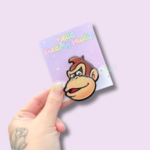 Puede incluir: Un pin de cara de mono de dibujos animados con cabeza marrón, cara bronceada y boca roja. El pin está en una tarjeta de color púrpura pastel con el texto "hello dreamy studio". El pin está sostenido por una mano.