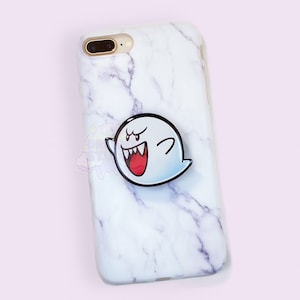 Puede incluir: Una funda de iPhone de mármol blanco con un agarre de teléfono con el personaje Boo. El personaje Boo es blanco con un contorno negro y tiene una boca roja y ojos negros.
