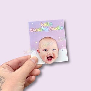 May include: A square card with a baby's face cut out, smiling widely. The card has a light purple background with the words "hello dreamy Studio" in rainbow colors. Small stars and clouds decorate the card.