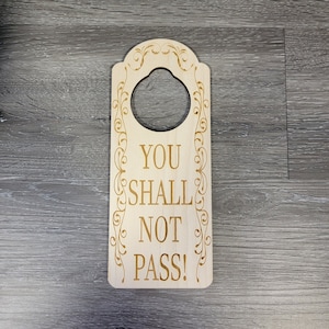 Puede incluir: Un colgador de puerta de madera con un borde decorativo y un agujero para colgar. El texto "YOU SHALL NOT PASS!" está grabado en dorado en el colgador.