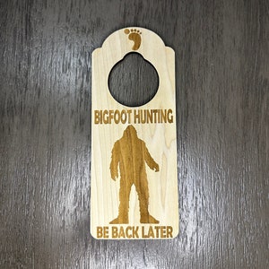 Könnte beinhalten: Hölzerner Türhänger mit einer Silhouette von Bigfoot und dem Text "BIGFOOT HUNTING BE BACK LATER".