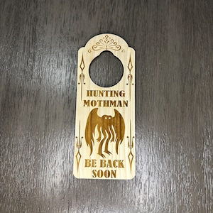 Könnte beinhalten: Hölzerner Türhänger mit einer braunen Silhouette einer Kreatur mit Flügeln und dem Text "Hunting Mothman Be Back Soon".