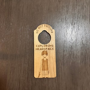 Puede incluir: Un colgador de puerta de madera con el texto "No molestar" y "Explorando el espacio mental" con una ilustración de dibujos animados de una persona.