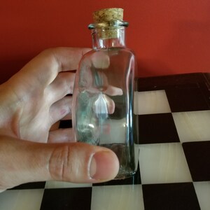May include: A clear glass bottle with a cork stopper. The bottle is empty and has a slightly rounded shape.