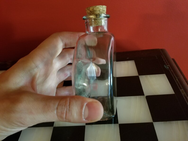 May include: A clear glass bottle with a cork stopper. The bottle is empty and has a slightly rounded shape.