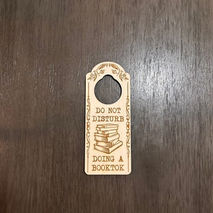 Könnte beinhalten: Ein hölzerner Türhänger mit dem Text "Nicht stören" und einem Stapel Bücher mit dem Text "Doing a Booktok".