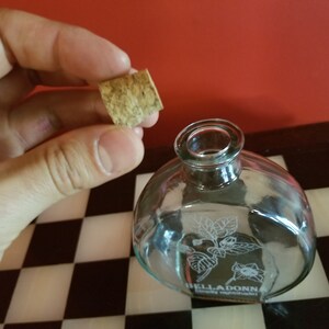 May include: A clear glass bottle with a cork stopper. The bottle has a floral design etched into the glass and the text "BELLADONNA" on the bottom. The bottle is sitting on a black and white checkered surface.