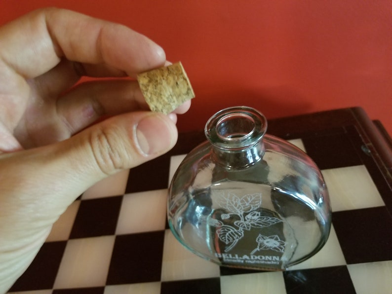 May include: A clear glass bottle with a cork stopper. The bottle has a floral design etched into the glass and the text "BELLADONNA" on the bottom. The bottle is sitting on a black and white checkered surface.