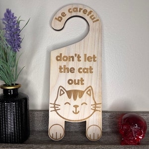Könnte beinhalten: Hölzerner Türhänger mit einer Katzenillustration und dem Text "be careful don't let the cat out".