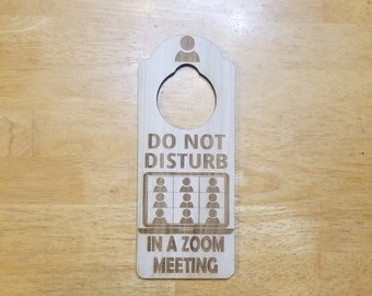 Puede incluir: Un colgador de puerta de madera con el texto "No molestar, estoy en una reunión de Zoom". El colgador tiene una ranura para una perilla de puerta y un gráfico de una pantalla de computadora con nueve personas en una videoconferencia.