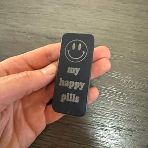 May include: A black rectangular object with rounded corners, featuring a smiling face and the words "my happy pills" engraved in a sans-serif font. The object is held in a hand.