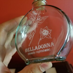 May include: A clear glass bottle with a cork stopper and a white etched design of a flower and leaves. The bottle is labeled "BELLADONNA (deadly nightshade)".