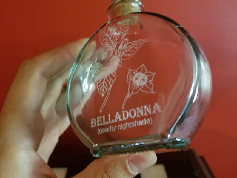 May include: A clear glass bottle with a cork stopper and a white etched design of a flower and leaves. The bottle is labeled "BELLADONNA (deadly nightshade)".