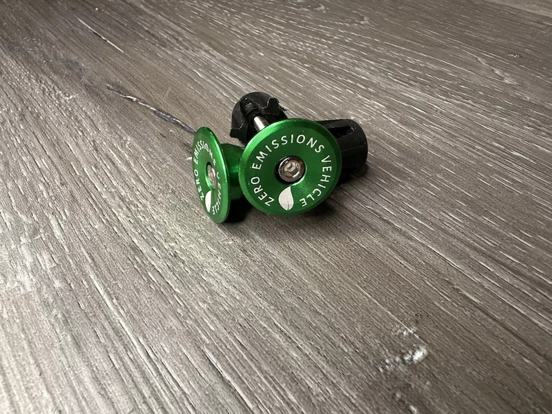 Puede incluir: Dos tapones de v&aacute;lvula de bicicleta verdes con tapas negras. El tap&oacute;n delantero tiene el texto "ZERO EMISSIONS VEHICLE" impreso.
