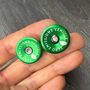 Puede incluir: Dos tapones de v&aacute;lvula redondos de color verde con el texto "ZERO EMISSIONS VEHICLE" y un peque&ntilde;o dise&ntilde;o de hoja.