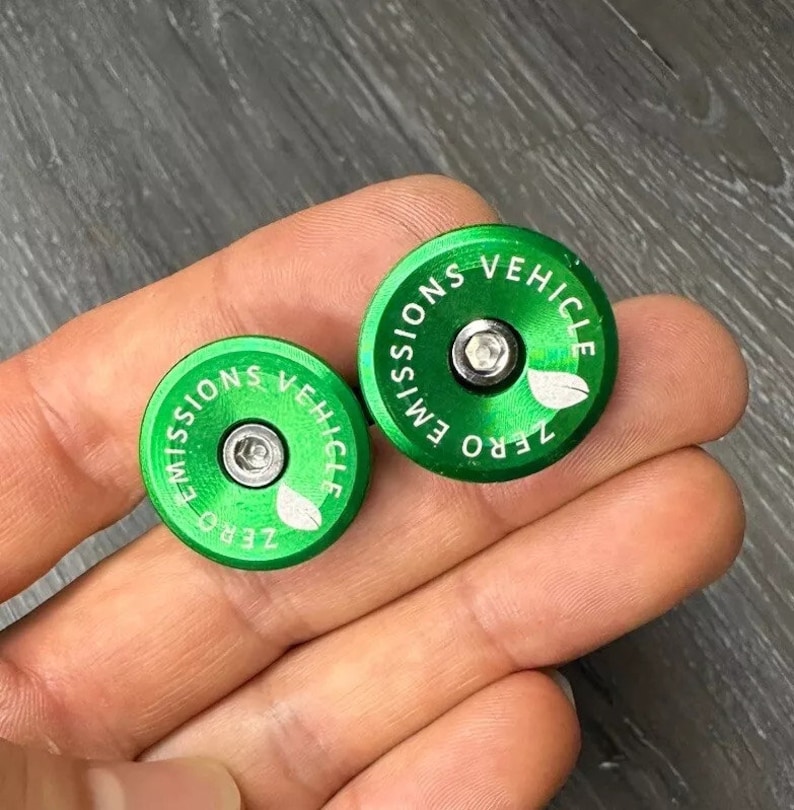 Puede incluir: Dos tapones de v&aacute;lvula redondos de color verde con el texto "ZERO EMISSIONS VEHICLE" y un peque&ntilde;o dise&ntilde;o de hoja.