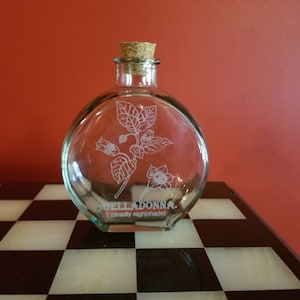 May include: A clear glass bottle with a cork stopper and an etched design of a belladonna plant. The bottle is labeled "BELLADONNA (deadly nightshade)".