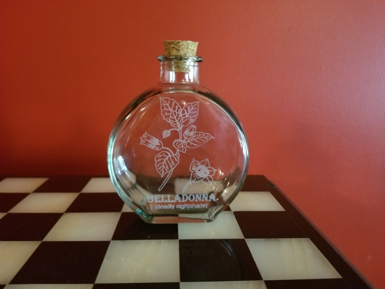 May include: A clear glass bottle with a cork stopper and an etched design of a belladonna plant. The bottle is labeled "BELLADONNA (deadly nightshade)".