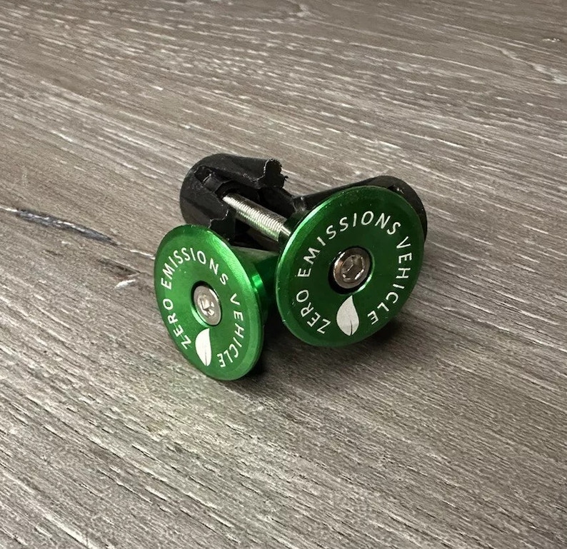 Puede incluir: Dos tapas de v&aacute;stago de bicicleta verdes con un tornillo negro. Las tapas tienen un dise&ntilde;o de hoja blanca y el texto "ZERO EMISSIONS VEHICLE".