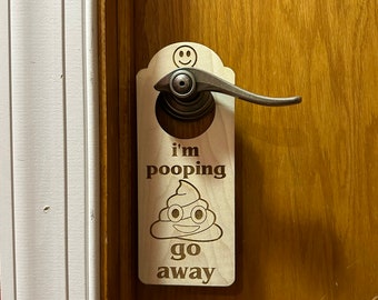 Puede incluir: Un colgador de puerta de madera con el texto "i'm pooping go away" y un emoji de caca de dibujos animados con una cara sonriente.