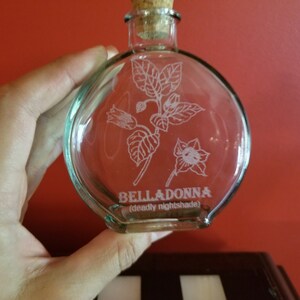 May include: A clear glass bottle with a cork stopper and a white etched design of a flower and leaves. The bottle is labeled "BELLADONNA (deadly nightshade)".