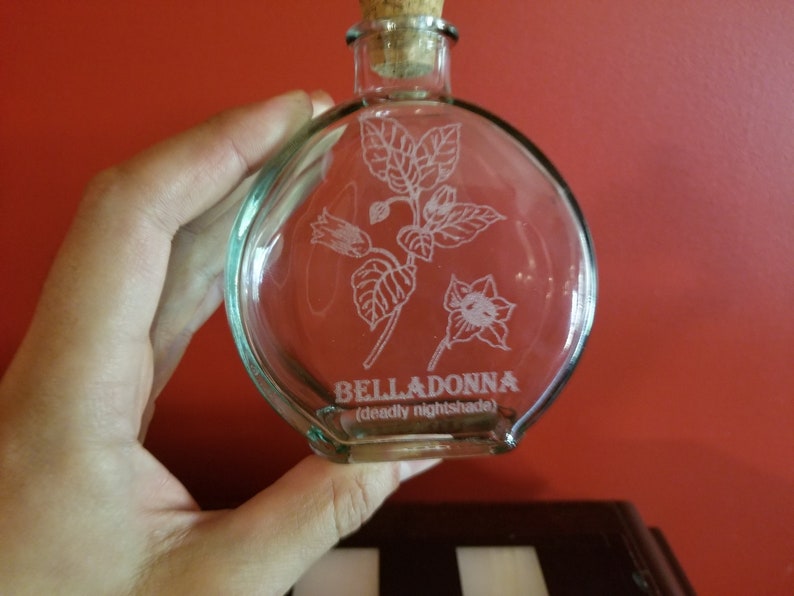 May include: A clear glass bottle with a cork stopper and a white etched design of a flower and leaves. The bottle is labeled "BELLADONNA (deadly nightshade)".