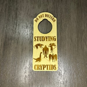 Könnte beinhalten: Hölzerner Türhänger mit dem Text "Nicht stören Studiere Kryptiden". Der Türhänger hat ein Loch für einen Türknauf und zeigt eine Silhouette von Bigfoot, einem geflügelten Wesen, einem Dinosaurier und anderen Kryptiden.