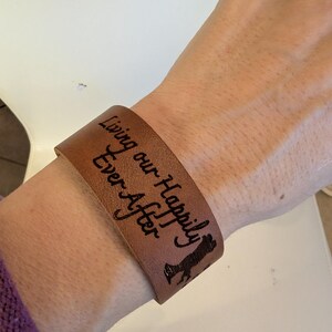May include: A brown leather cuff bracelet with the words "Living our Happily Ever After" engraved in black. A silhouette of a couple is also engraved on the bracelet. The bracelet is worn on a wrist.