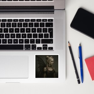 Peut inclure: Un autocollant avec une image d'art numérique d'une fée. La fée a des oreilles pointues, des ailes sombres et une robe sombre. Elle a la peau pâle et des cheveux blonds en deux chignons. Le fond est vert foncé.