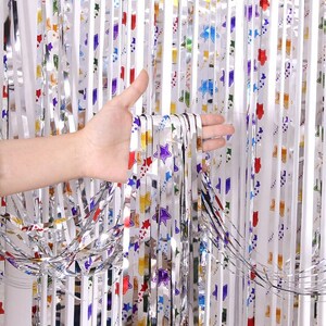Pode incluir: Cortina de franjas metálicas prateadas com padrões coloridos de estrelas e formas. A cortina é segurada por uma mão, mostrando o material brilhante e refletor. Ideal para decorações de festas e eventos festivos.