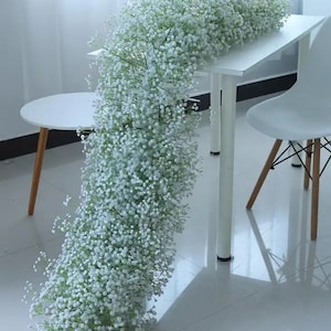 May include: A long garland of white baby's breath flowers draped over a white table with a white chair. The garland is made of small, delicate flowers and is perfect for adding a touch of elegance to any event.