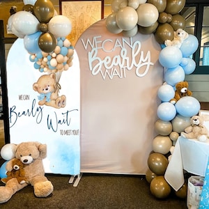 May include: A baby shower backdrop with a teddy bear theme. The backdrop features a sign with the text "We Can Bearly Wait To Meet You!" and a large balloon arch in shades of brown, white, and blue. Teddy bears are placed throughout the display.