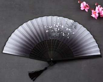 Puede incluir: Un abanico plegable negro y gris con un diseño floral blanco. El abanico tiene un mango negro y una borla.