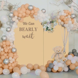 May include: A baby shower backdrop featuring a beige arch of balloons, silver orbs, and white and peach balloons. The backdrop has the text "We Can Bearly Wait" and a teddy bear.