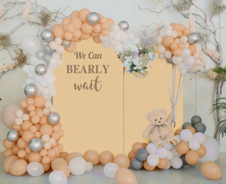 May include: A baby shower backdrop featuring a beige arch of balloons, silver orbs, and white and peach balloons. The backdrop has the text "We Can Bearly Wait" and a teddy bear.