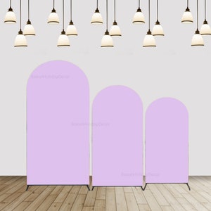 May include: Three different sizes of light purple arch backdrops for photography. The backdrops are made of fabric and are standing on black metal stands. The backdrops are in front of a white wall with white hanging lights.