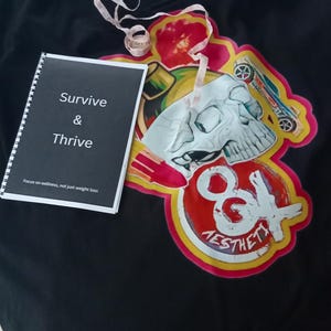 Puede incluir: Camiseta negra con un diseño gráfico colorido que presenta una calavera, una bomba y el texto "8& Aesthetic". Un cuaderno blanco con el texto "Survive & Thrive Focus on wellness, not just weight loss" está encima de la camiseta.