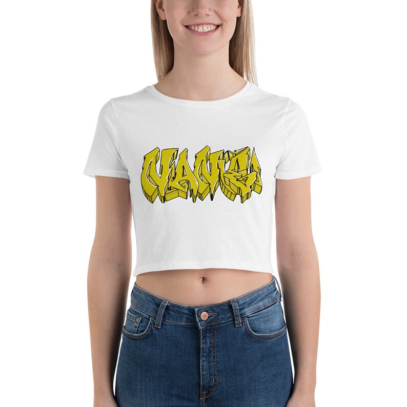 May include: White crop top with the word "WAVE" in yellow graffiti-style lettering. The shirt is short-sleeved and made of a soft material. The model is wearing blue jeans.