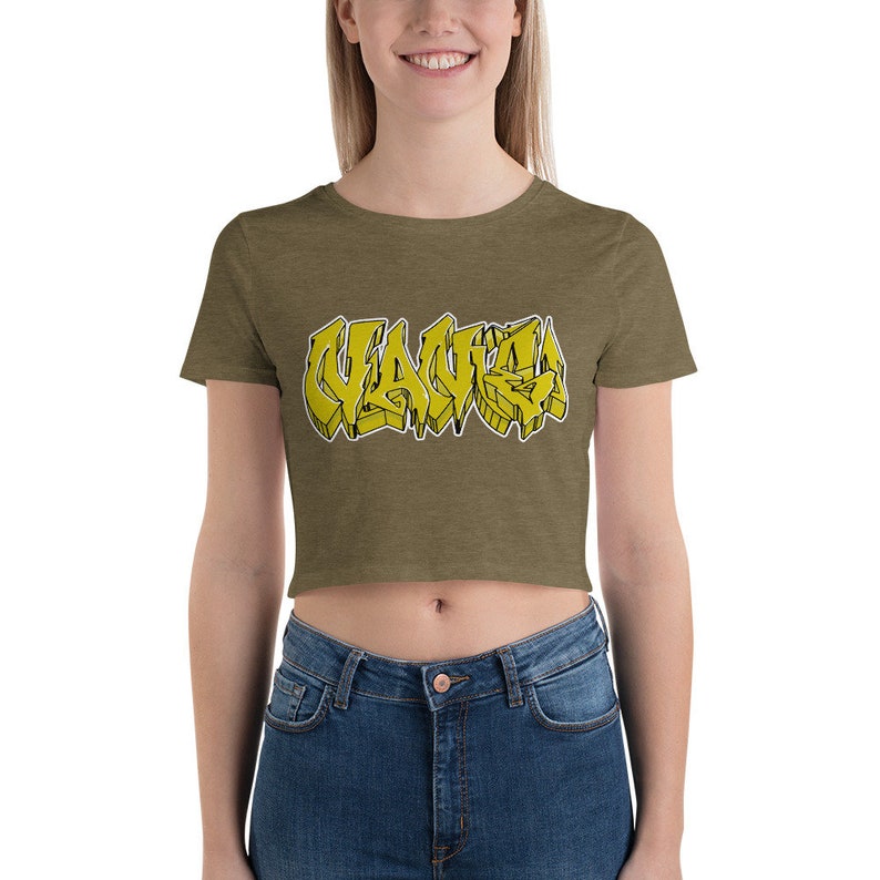 May include: Olive green crop top with the word "VAUVA" in yellow graffiti-style lettering. The text has a black outline. The shirt is made of a soft material and is paired with blue jeans.