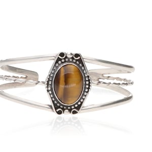 May include: A silver cuff bracelet featuring an oval tiger's eye stone at the center. The stone is surrounded by a decorative bezel. The bracelet has a three-band design with a twisted rope detail.