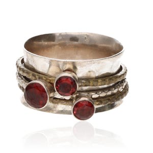 May include: A silver-toned ring with three red gemstones. The ring has a wide band with a hammered texture and several smaller bands with textured and smooth surfaces. The gemstones are round and set in silver bezels.