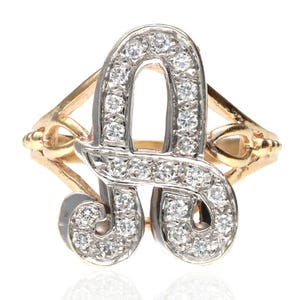 以下が含まれることがあります： 小さなダイヤモンドがちりばめられた、金色の「A」の文字が特徴の指輪。 文字は銀色の金属で縁取られ、バンドは装飾的な透かし彫りのデザインです。 ジュエリー。