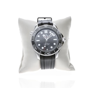 May include: A black and silver wristwatch with a black rubber band. The watch face is dark gray with wave-like patterns and white markers. The watch is displayed on a white pillow.