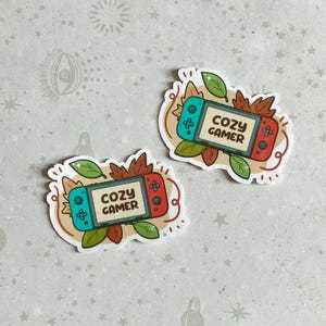 May include: Two stickers featuring a cartoon illustration of a Nintendo Switch console with autumn leaves and the text "Cozy Gamer".