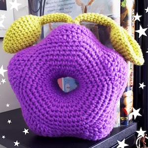 Könnte beinhalten: Ein violettes, gehäkeltes Kissen in Sternform mit zwei gelben, gehäkelten Griffen. Das Kissen liegt auf einem schwarzen Regal.