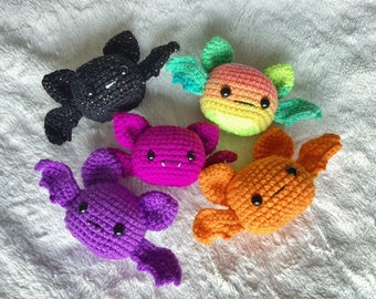 May include: Five crocheted stuffed animals in the shapes of bats and a dragon. The bats are black, purple, and pink. The dragon is orange and has rainbow wings.
